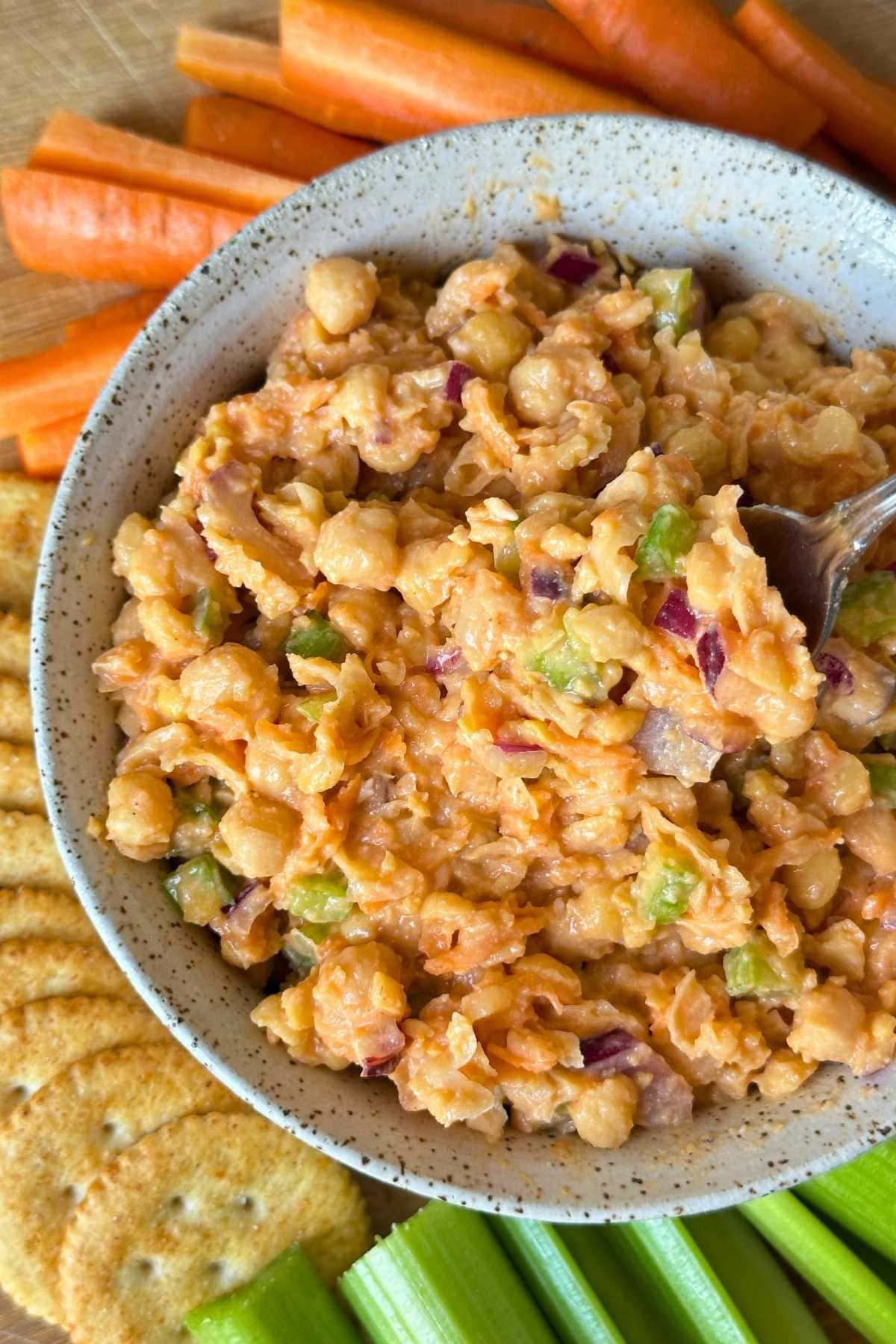 Smashed buffalo chickpea salad combined in a ceramic bowl surrounded by carrot sticks, crackers, and celery sticks.