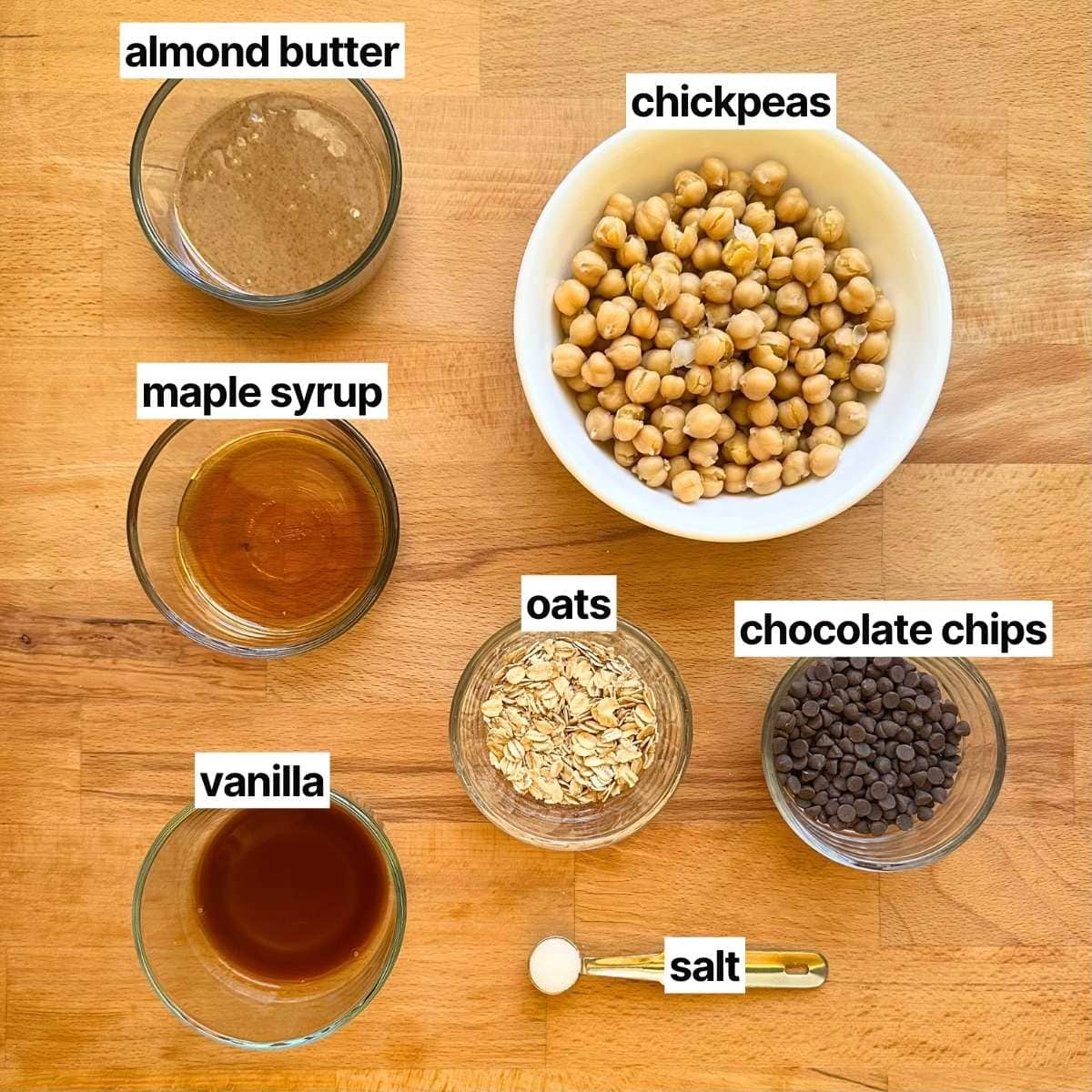 Ingredients to make chickpea cookie dough with corresponding labels: almond butter, chickpeas, vanilla, rolled oats, chocolate, maple syrup, and sea salt.