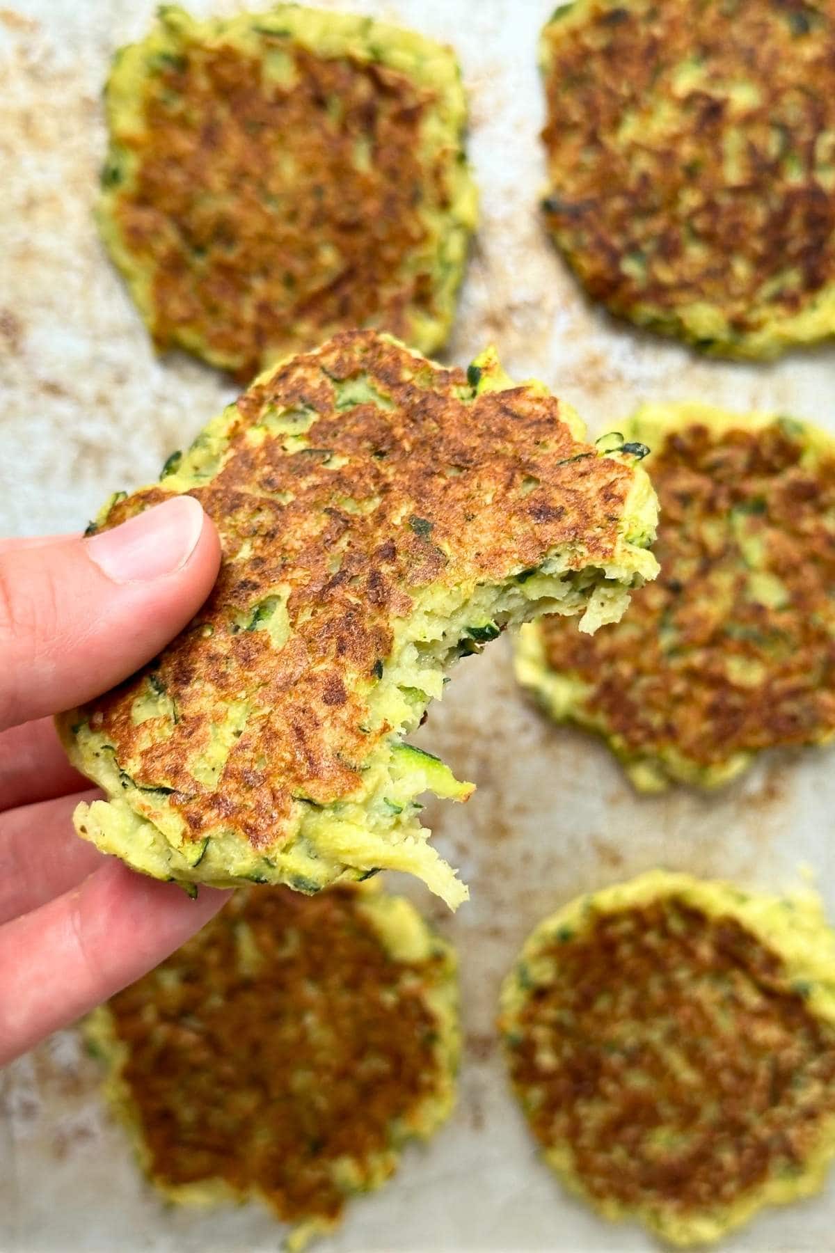Hand holding up a chickpea flour zucchini fritter with a bite taken out of it, more fritters are in the background on a baking sheet.