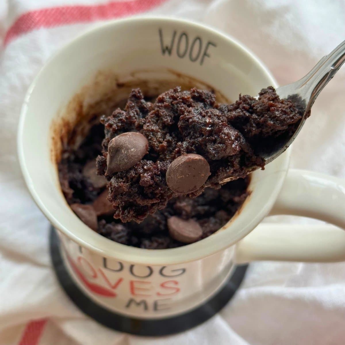 Fork with a bite of chocolate zucchini mug cake with chocolate chips on it, the chocolate mug cake is in the background on a white napkin.