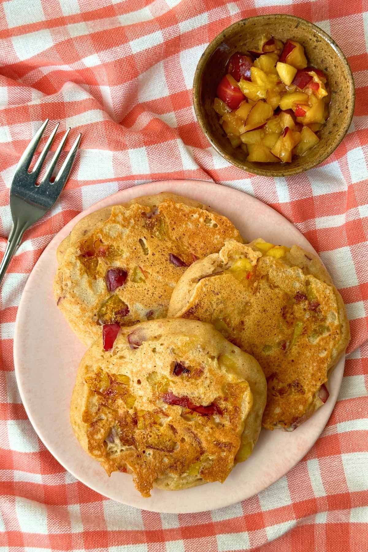 Three summer fruit pancakes on a plate over top a gingham napkin alongside a bowl of freshly chopped summer fruit.