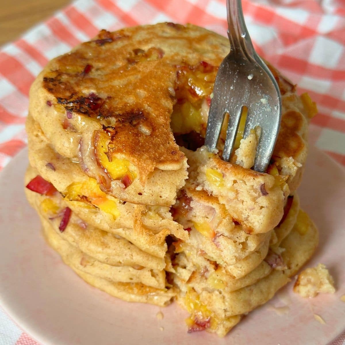 Stack of summer fruit pancakes with peaches sliced into and being eaten with a fork.