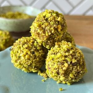 3 pistachio date balls mounded on a plate with more date bites and ground pistachio in the background.