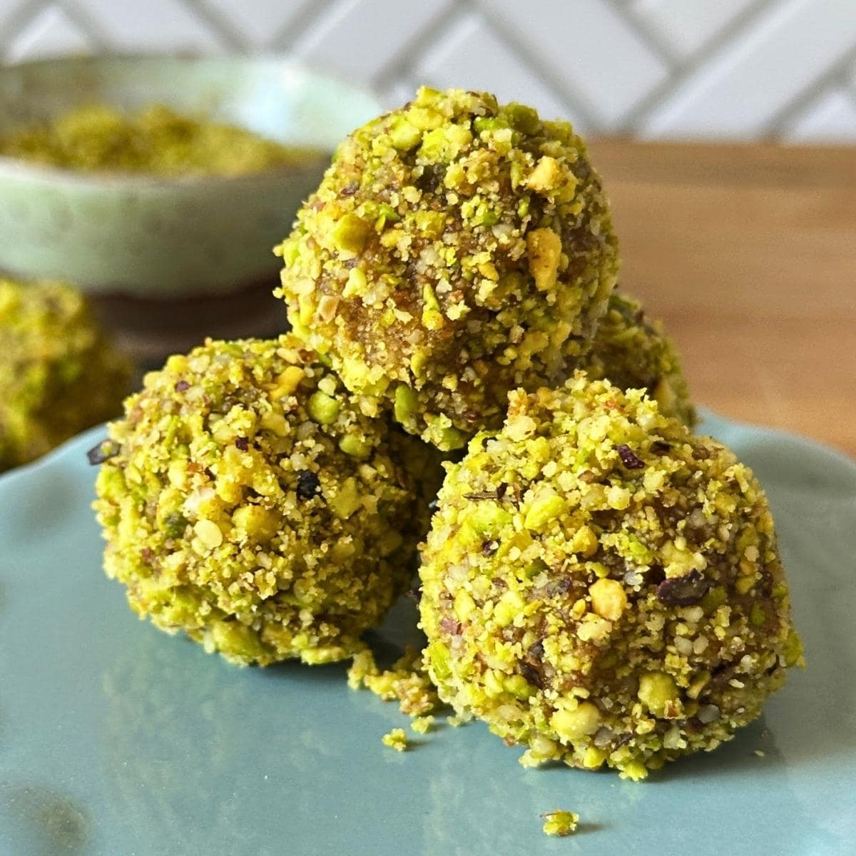 3 pistachio date balls mounded on a plate with more date bites and ground pistachio in the background.