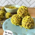 3 pistachio date balls mounded on a plate with more date bites and ground pistachio in the background.