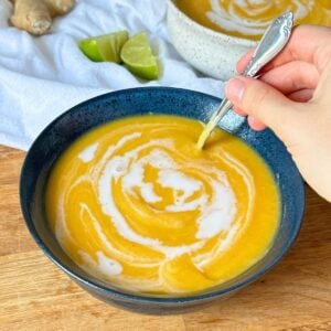 Creamy butternut squash soup with a swirl of coconut milk on top, another bowl of soup is in the background along with lime wedges and ginger.