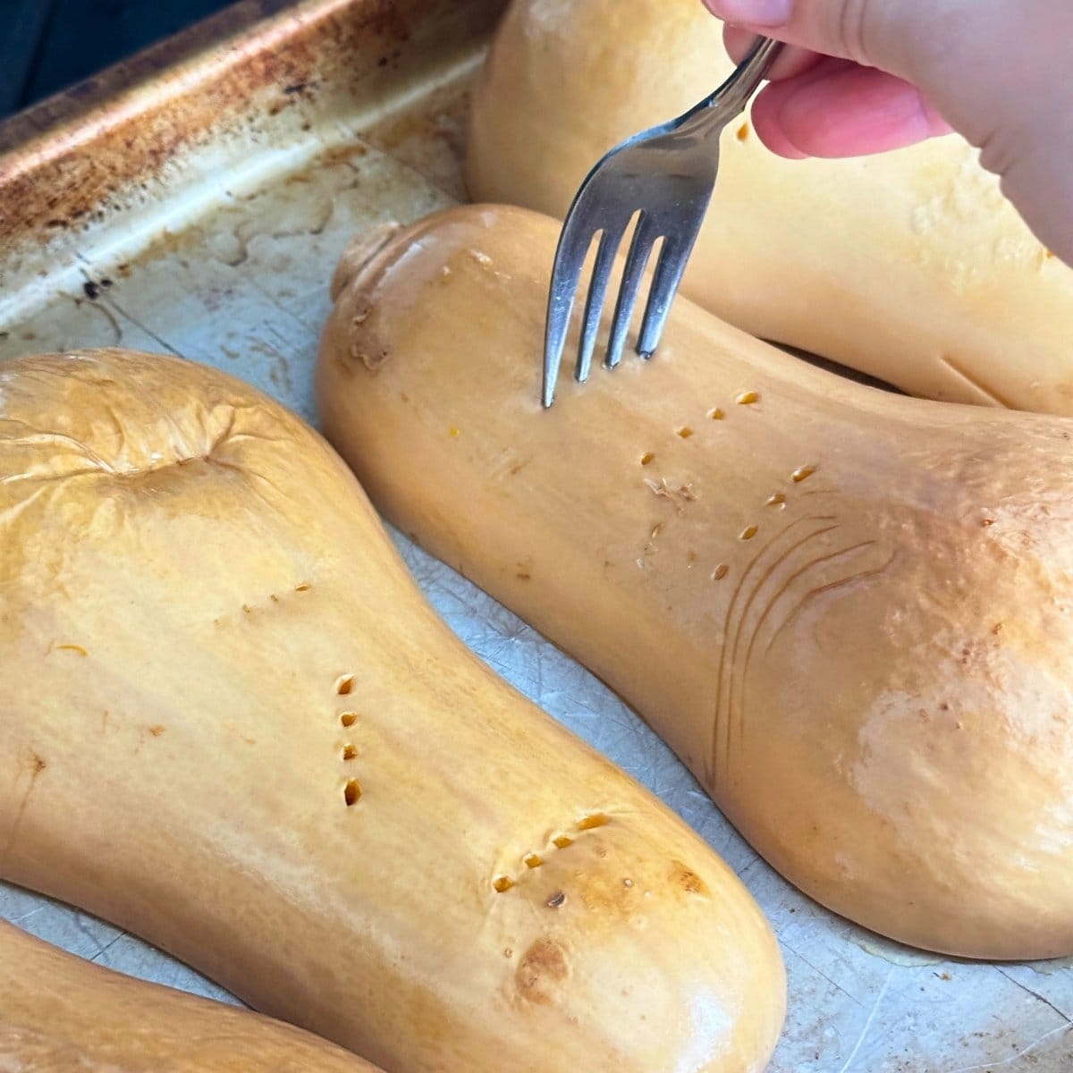Butternut squash halves cut side down on a baking sheet, pierced with a fork to test if they are fully cooked.