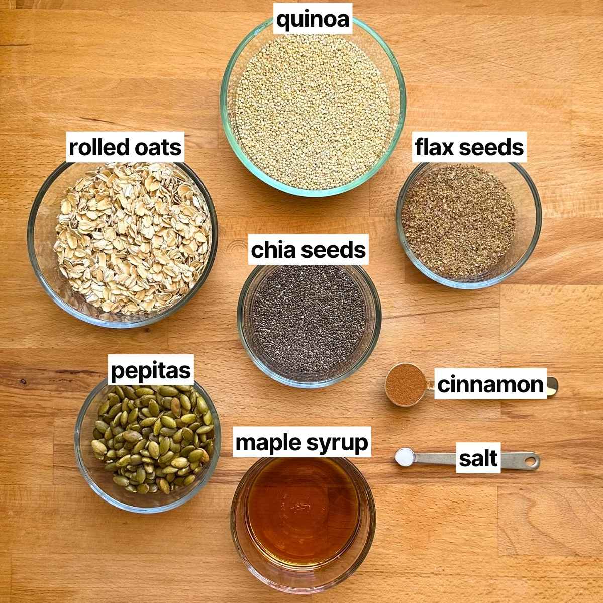 Ingredients needed to make popped quinoa granola with corresponding labels: quinoa, rolled oats, flax seeds, chia seeds, pepitas, maple syrup, cinnamon, and salt.