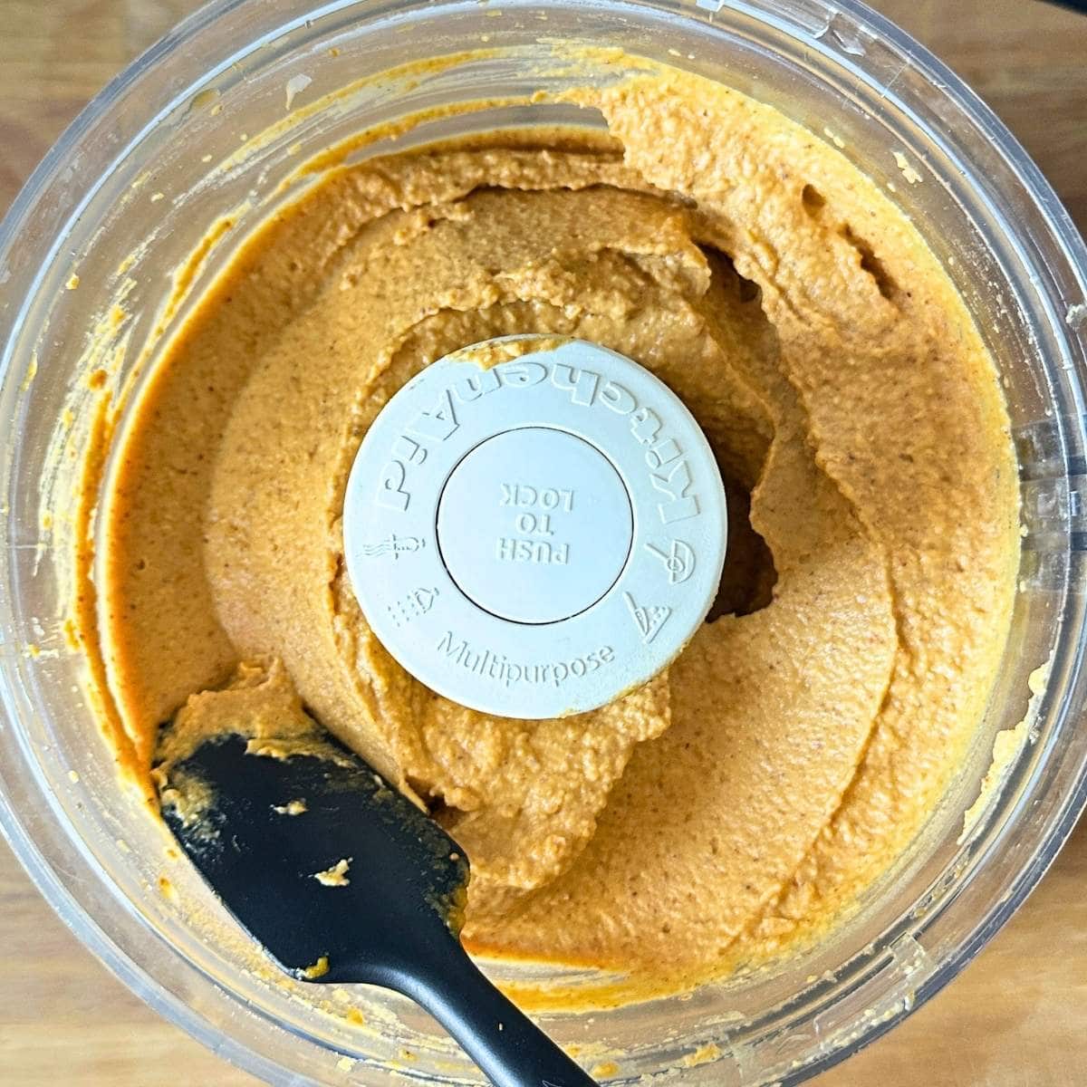 Pumpkin chickpea mixture in a food processor, with a spatula scraping down the smooth, creamy batter.