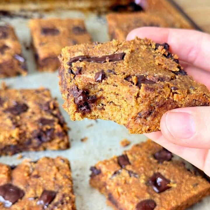 Pumpkin chickpea blondie with a bit taken out of it with lots of chocolate chunks, more vegan blondies are in the background on a baking sheet.