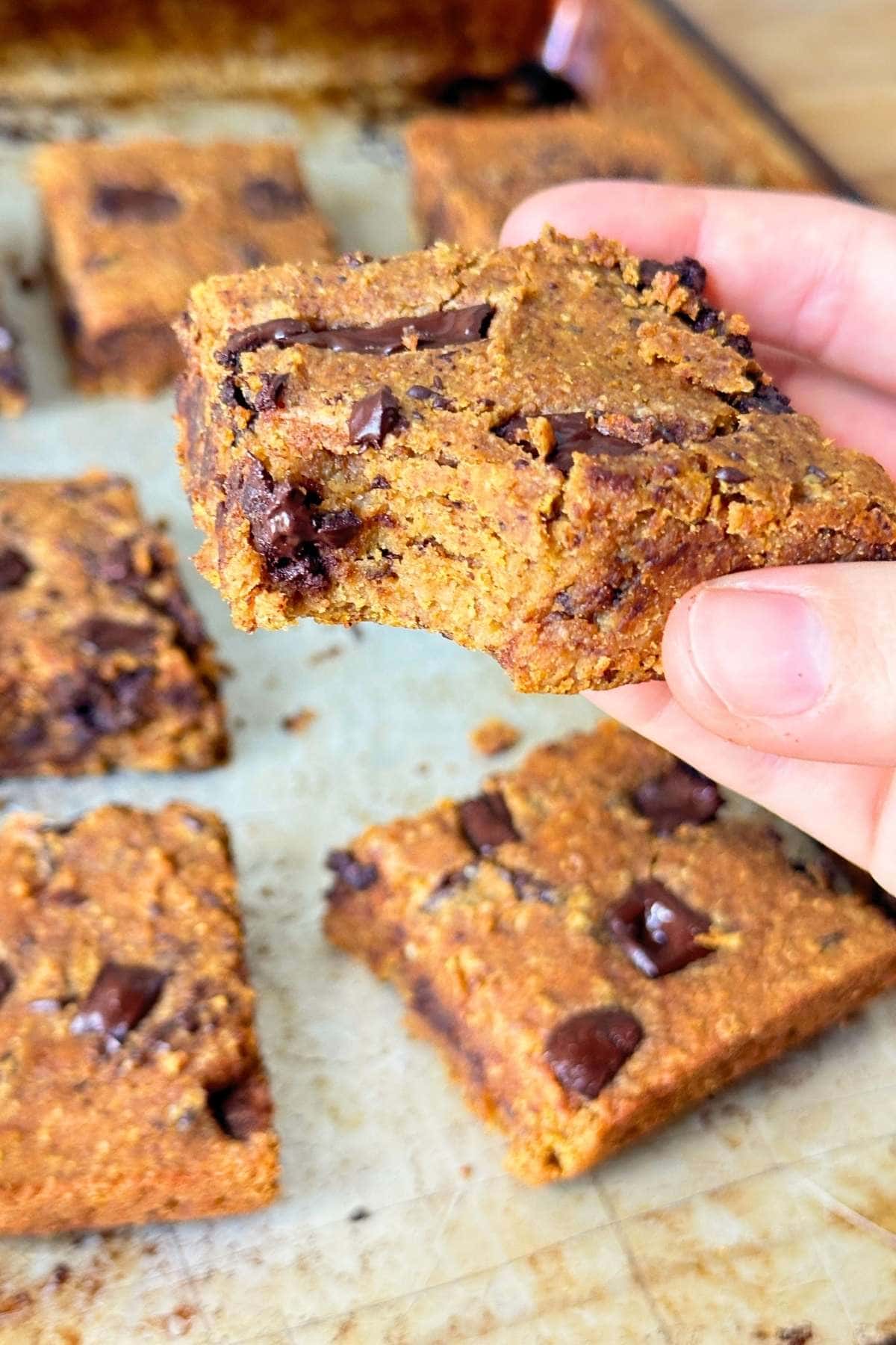 Hand holding a square of pumpkin chickpea blondie with chocolate chunks with a bite taken out of it.