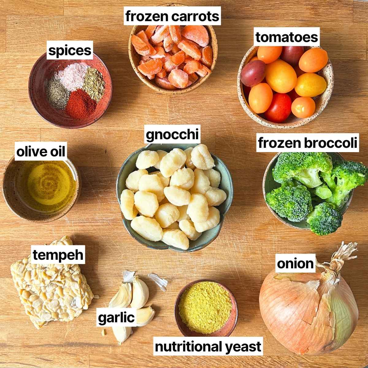 Ingredients needed to make sheet pan gnocchi with labels: spices, frozen carrots, tomatoes, olive oil, gnocchi, frozen broccoli, tempeh, garlic, nutritional yeast, and onion.
