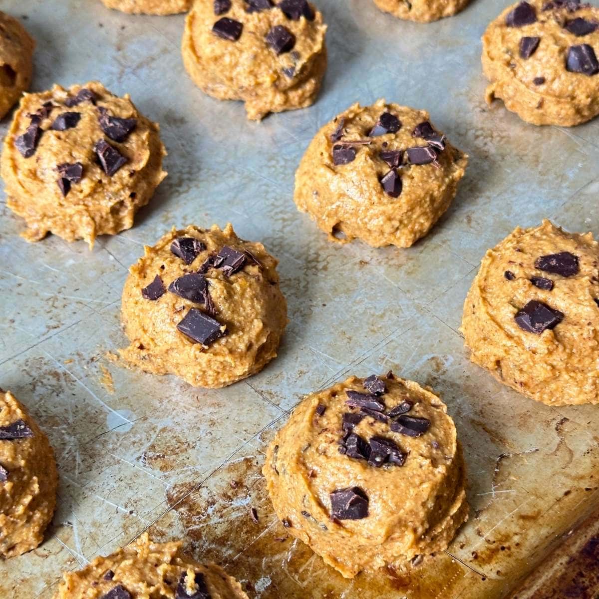 Chocolate chunks on top of unbaked pumpkin cookies on a baking sheet.