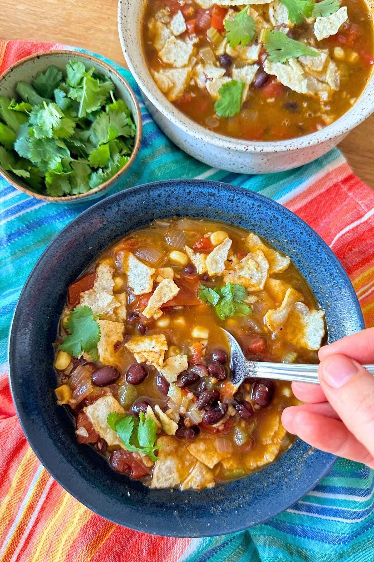 Vegan black bean tortilla soup in a blue bowl with a spoon over a bright red, orange, and blue napkin.
