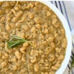 Bowl of creamy braised white beans with a sprig of rosemary on top.