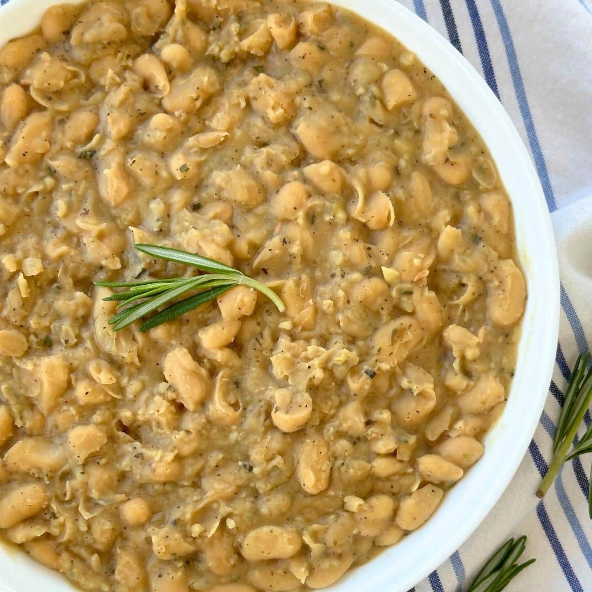 Braised white beans in a bowl, topped with a sprig of rosemary.