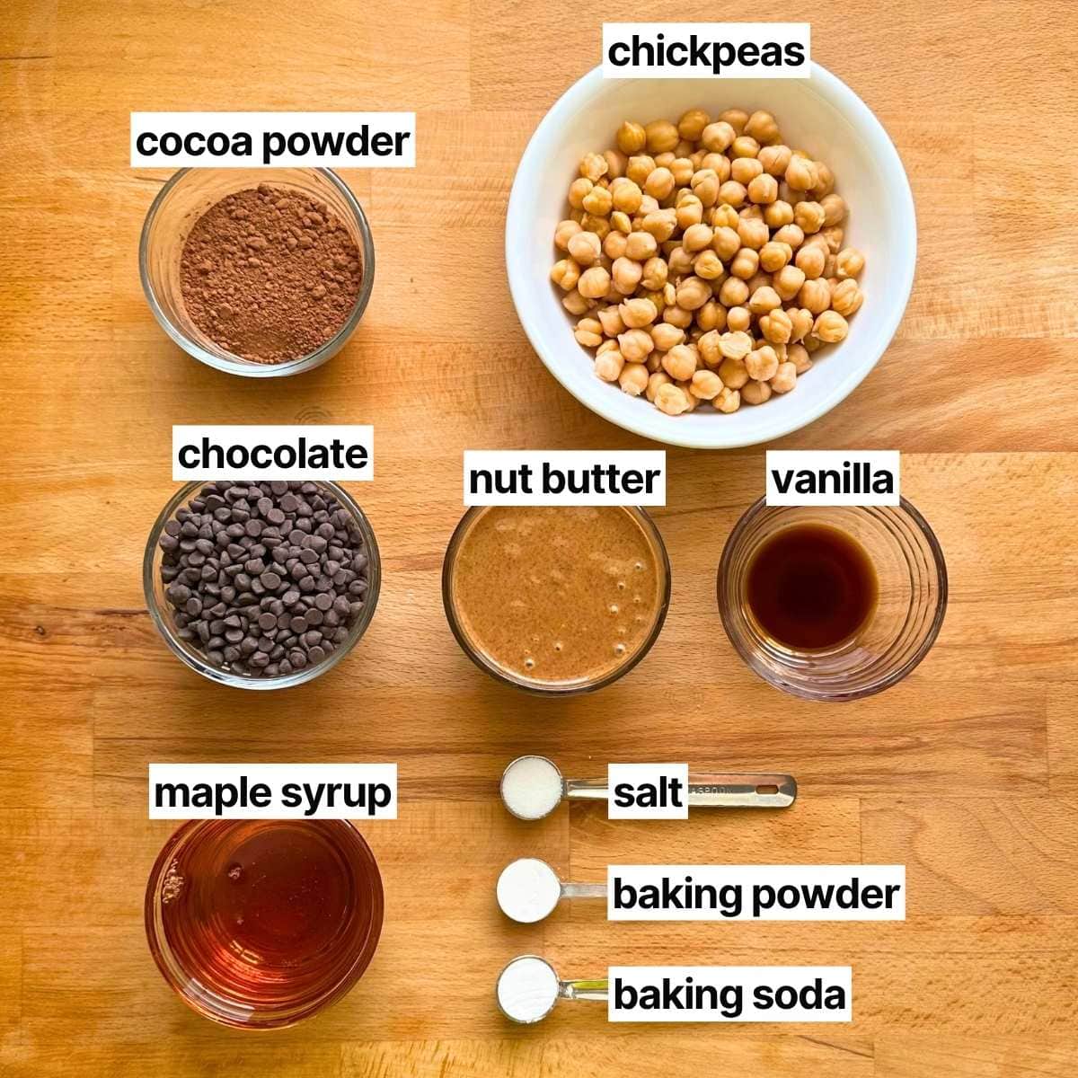 Ingredients to make chickpea brownies with corresponding labels: cocoa powder, chickpeas, chocolate, nut butter, vanilla, maple syrup, sea salt, baking powder, and baking soda.