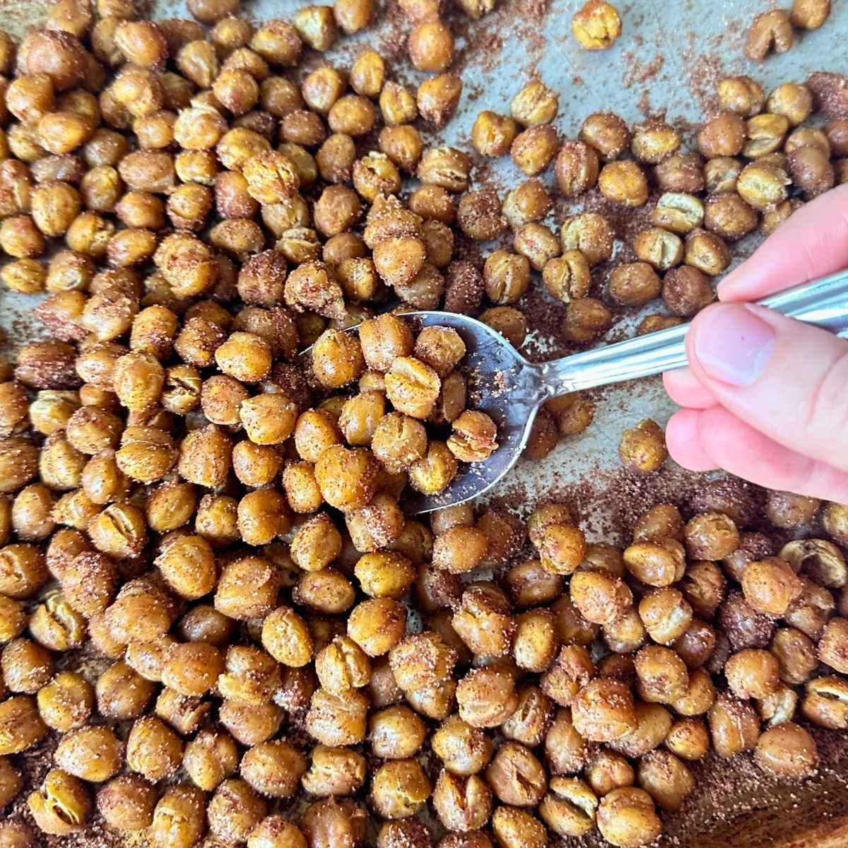 Cinnamon sugar chickpeas on a sheet pan with a large spoon scooping up the crunchy chickpeas coated in cinnamon sugar.