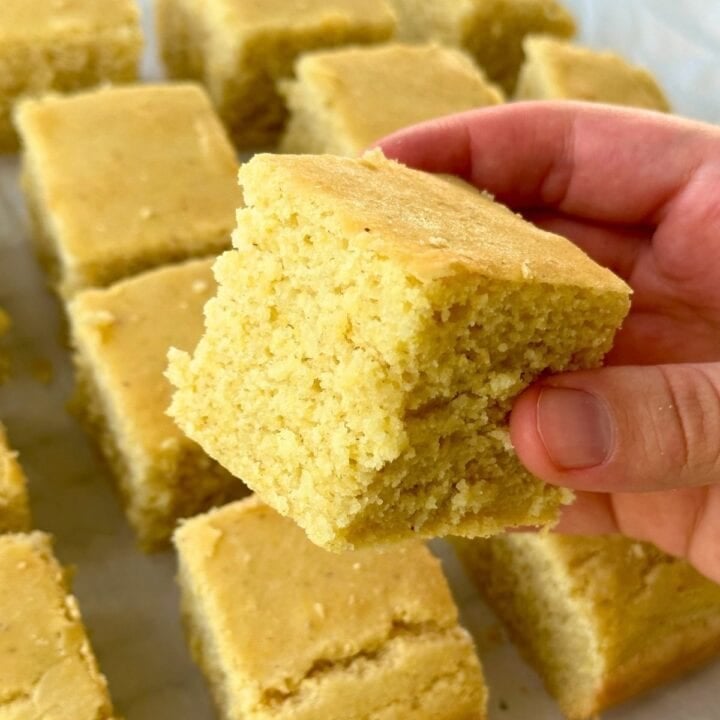 Cube of vegan gluten-free cornbread being held by a hand with more cornbread in the background.