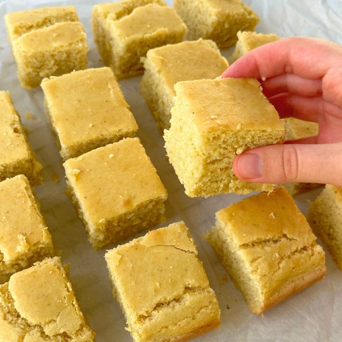Hand grabbing a square of gluten free vegan cornbread from a parchment sheet with more cornbread squares.