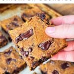 Hand holding a pumpkin chickpea blondie with big dark chocolate chunks, more blondies are in the background on a baking sheet.
