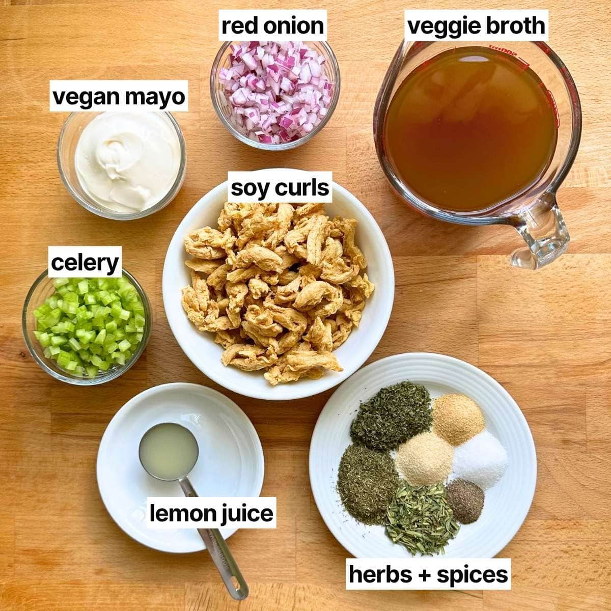 Ingredients to make vegan chicken salad with corresponding written labels: vegan mayo, red onion, veggie broth, celery, soy curls, lemon juice, herbs, and spices.