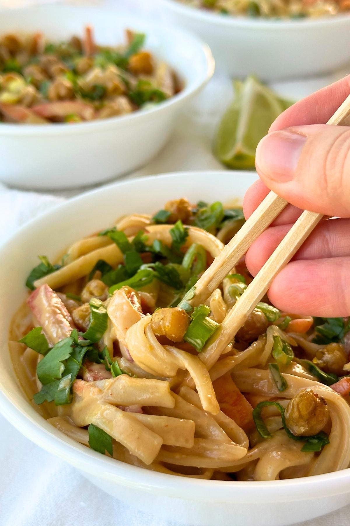 Chopsticks in a bowl of creamy vegan Thai peanut noodles topped with green onion, cilantro, and chickpeas.