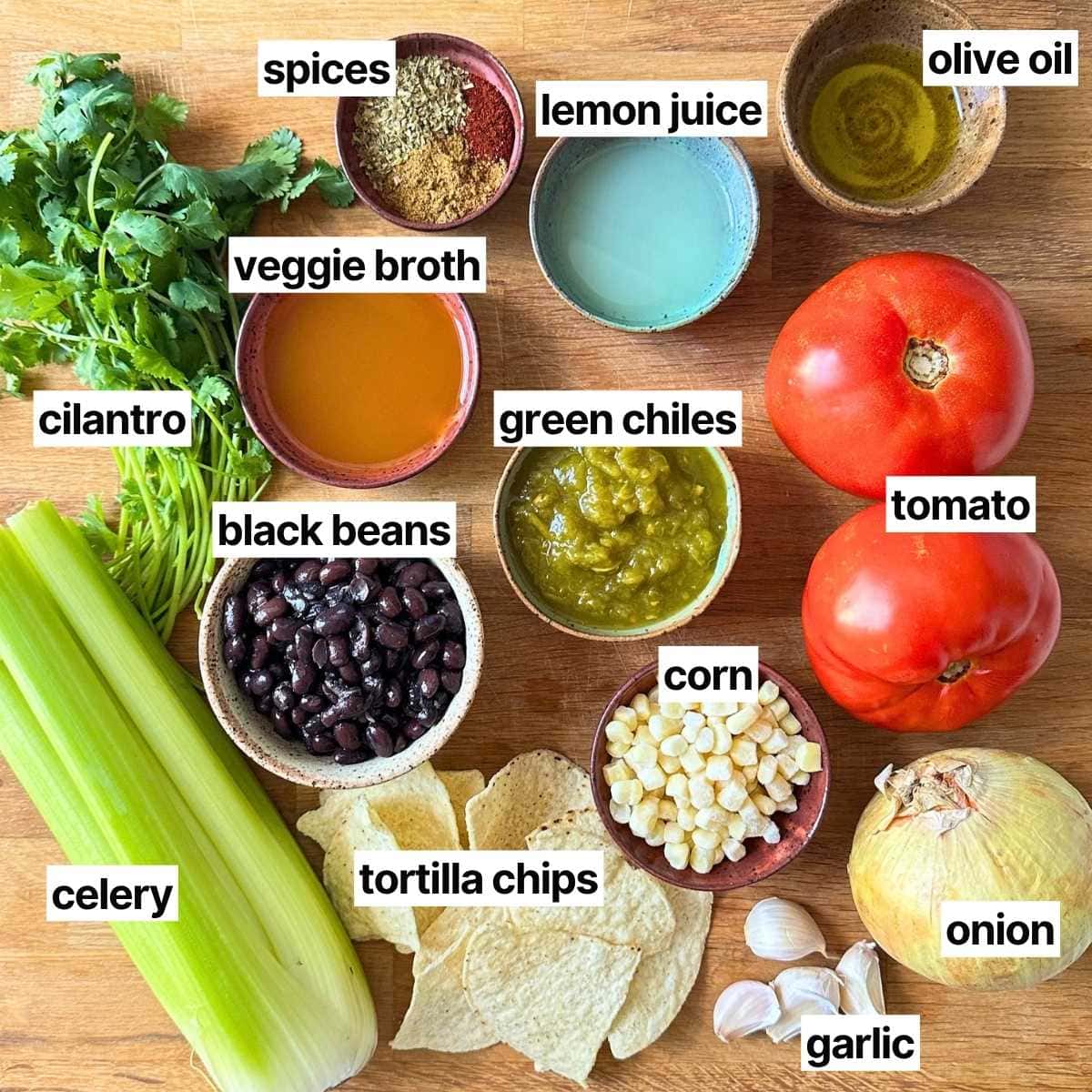Ingredients needed to make vegan black bean tortilla soup with labels: spices, lemon juice, olive oil, veggie broth, green chiles, tomato, cilantro, black beans, corn, celery, tortilla chips, garlic, and onion.