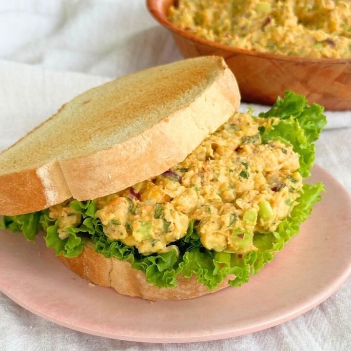 Chickpea chicken salad on a piece of bread with lettuce. Top piece of bread is slightly to the side. A bowl of vegan chicken salad is in the background.
