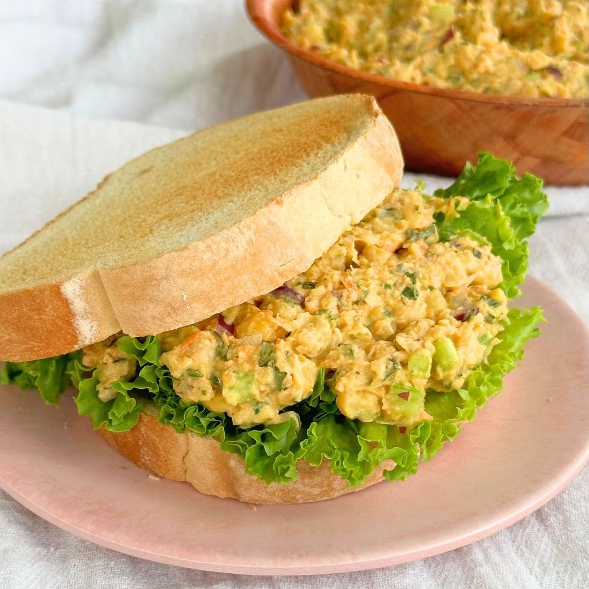 Chickpea chicken salad on a piece of bread with lettuce, top piece of bread is slightly to the side and a bowl of vegan chicken salad is in the background.
