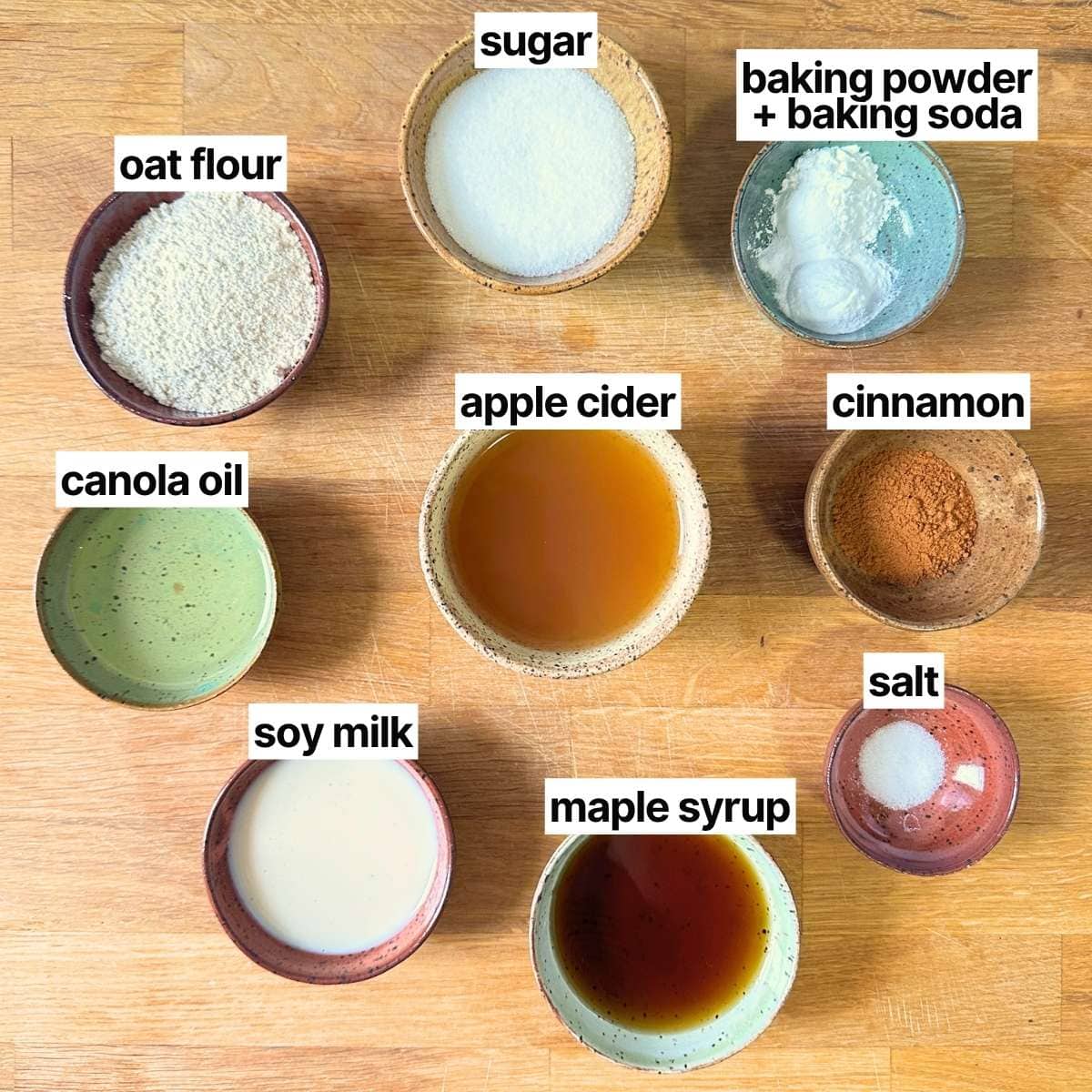 Ingredients needed to make vegan gluten free apple cider donuts with labels: oat flour, sugar, baking powder + baking soda, canola oil, apple cider, cinnamon, soy milk, maple syrup, and salt.