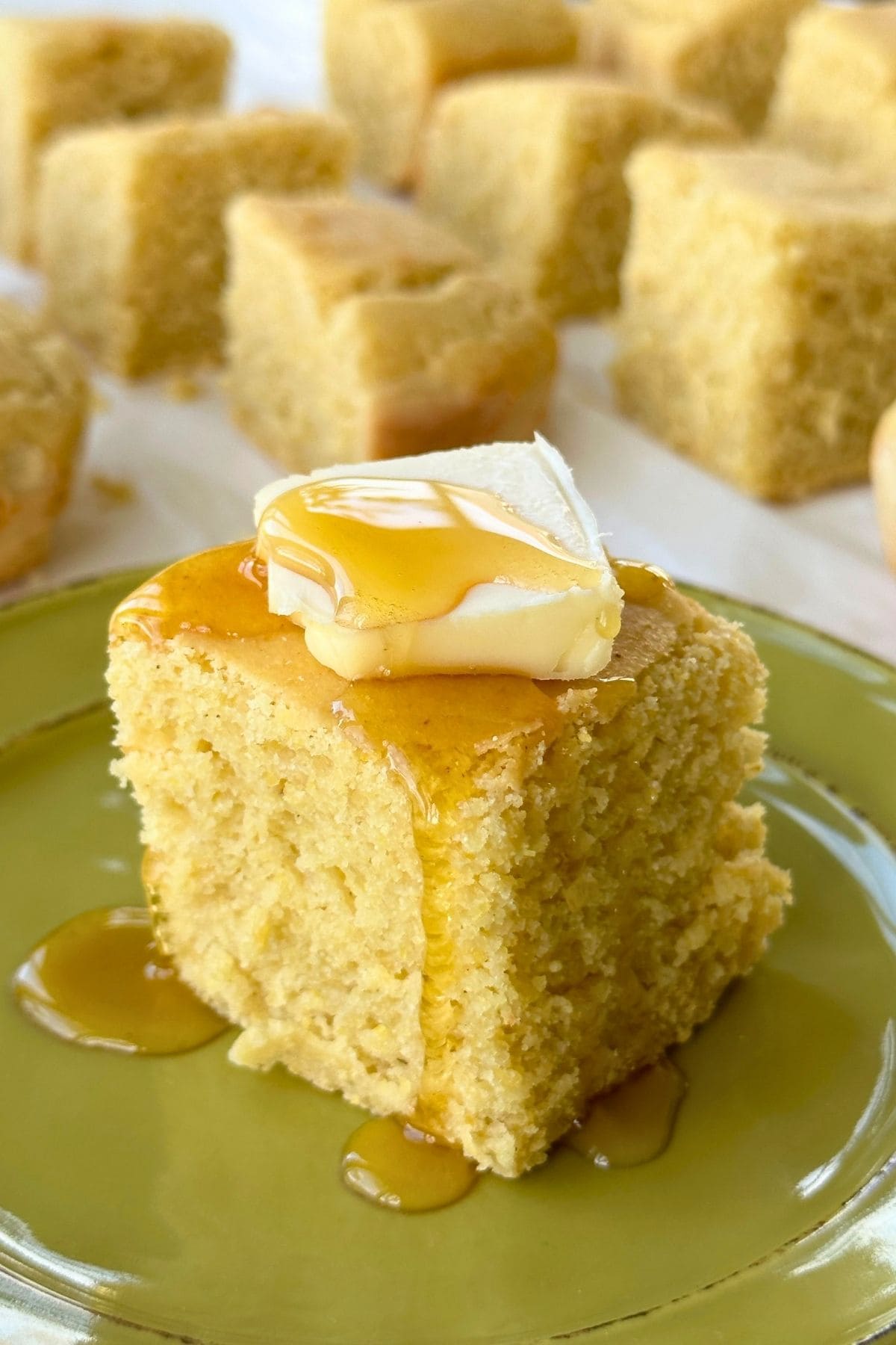 Cube of vegan gluten-free cornbread with butter and maple syrup dripping onto a green plate.