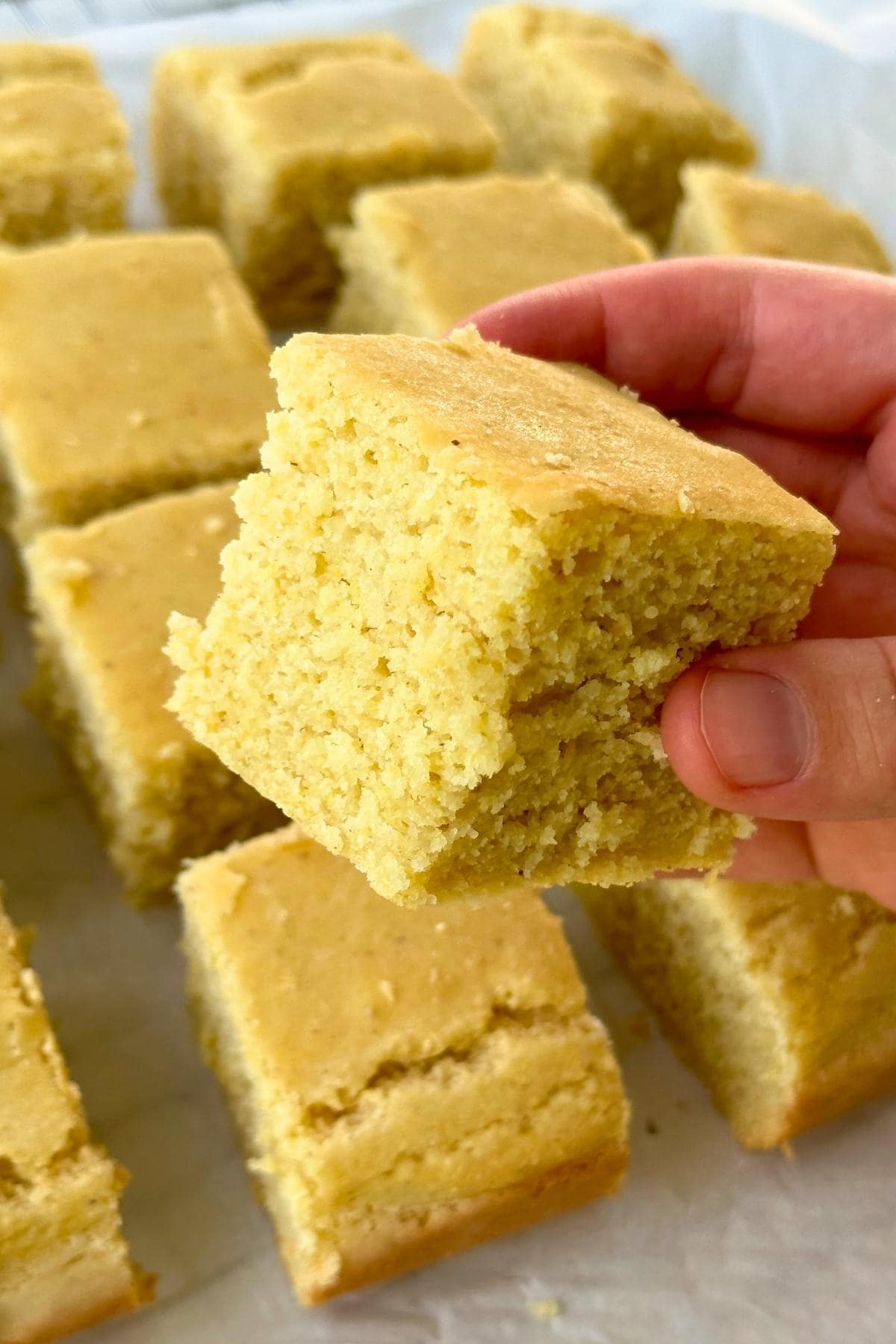 Cube of vegan gluten-free cornbread being held by a hand with more cornbread in the background.