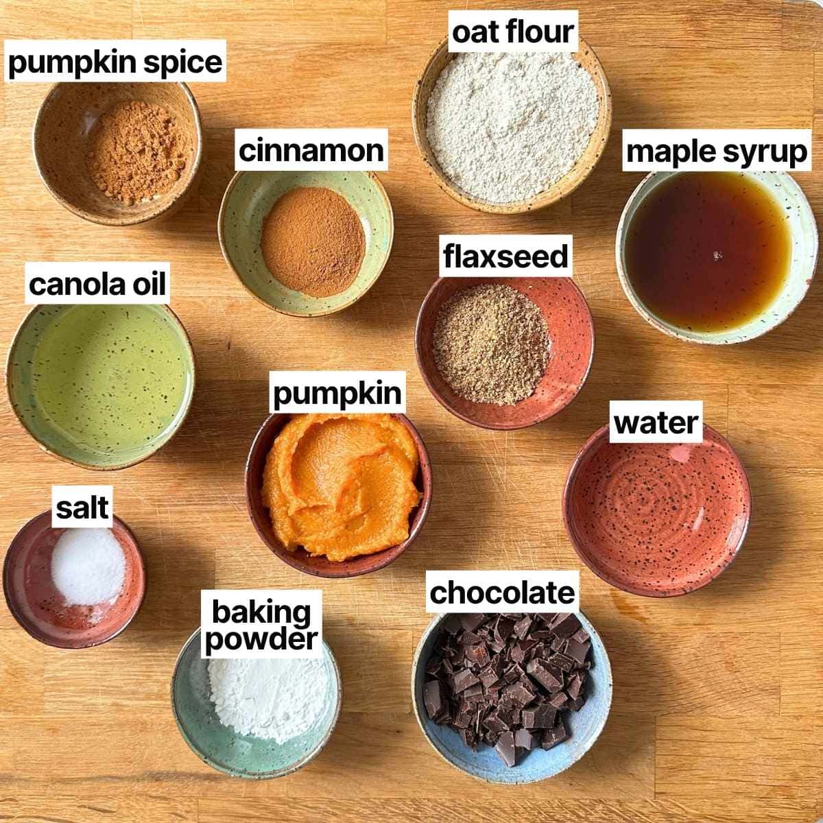 Ingredients needed to make vegan gluten-free pumpkin cookies with labels: pumpkin spice, cinnamon, oat flour, maple syrup, canola oil, pumpkin, flaxseed, water, salt, baking powder, and chocolate.