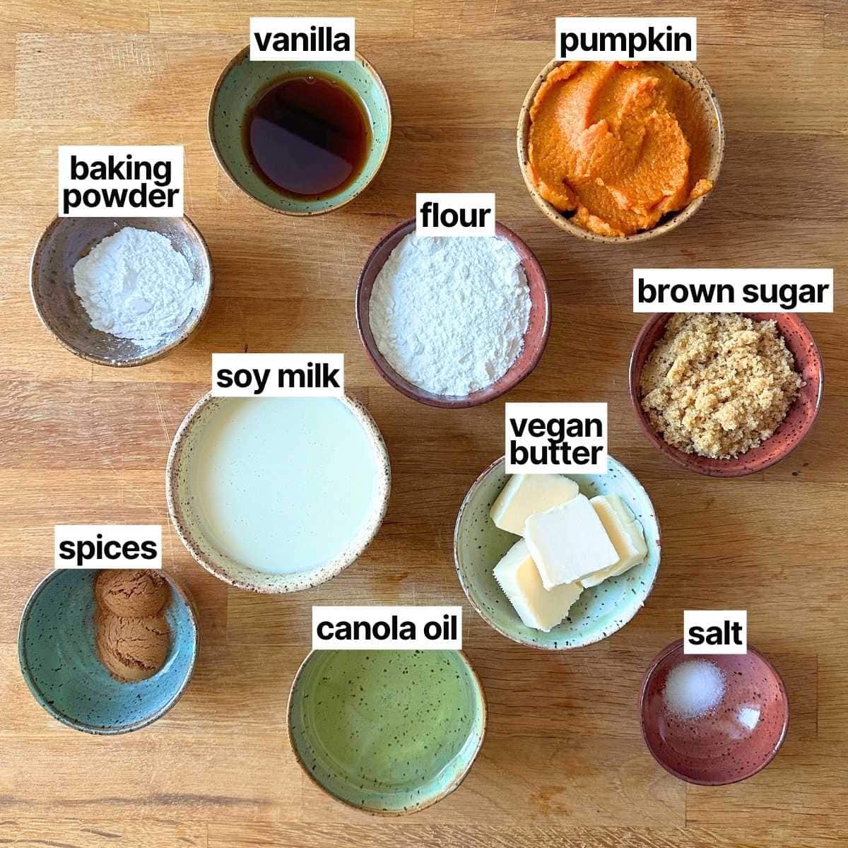 Ingredients needed to make vegan pumpkin muffins with brown sugar streusel with labels: baking powder, vanilla, pumpkin, flour, soy milk, brown sugar, vegan butter, spices, canola oil, and salt.