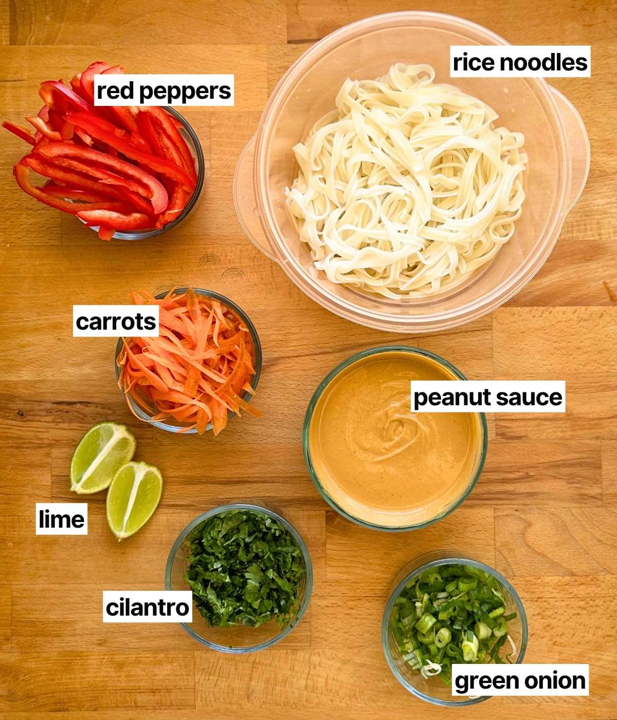 Ingredients needed to make peanut noodles with corresponding labels: rice noodles, peppers, carrots, lime wedges, peanut sauce, cilantro, and green onions.
