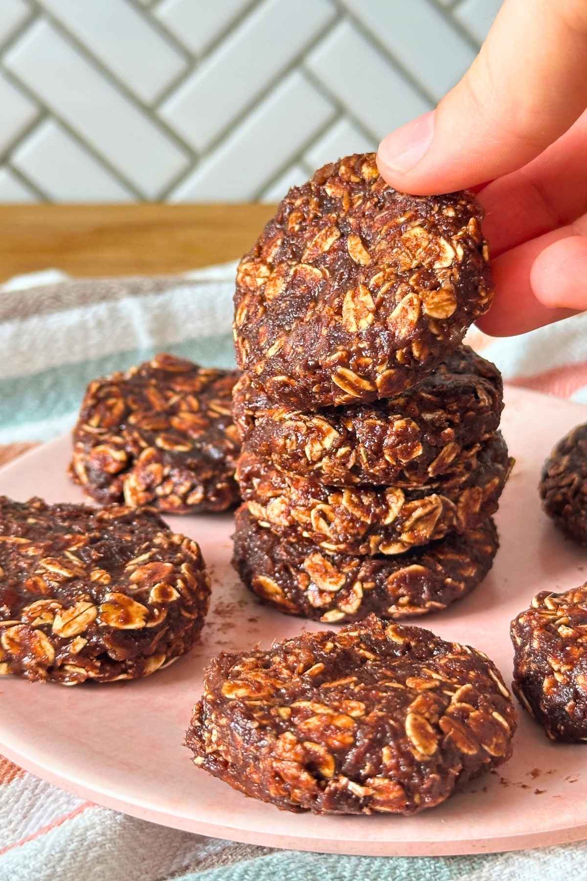 Hand picking up a no-bake date cookie from a stack of 4 cookies with more surrounding them on a pink plate.