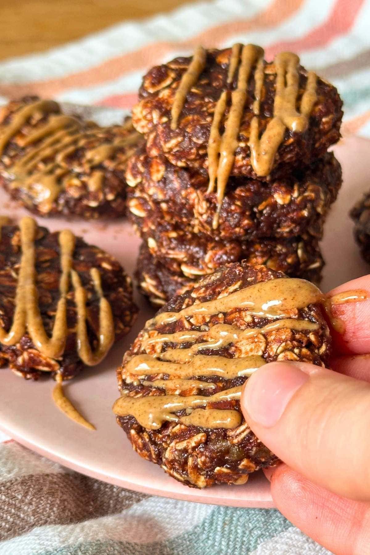 Hand grabbing a no-bake date cookie with almond butter drizzled on top from a plate with more cookies.