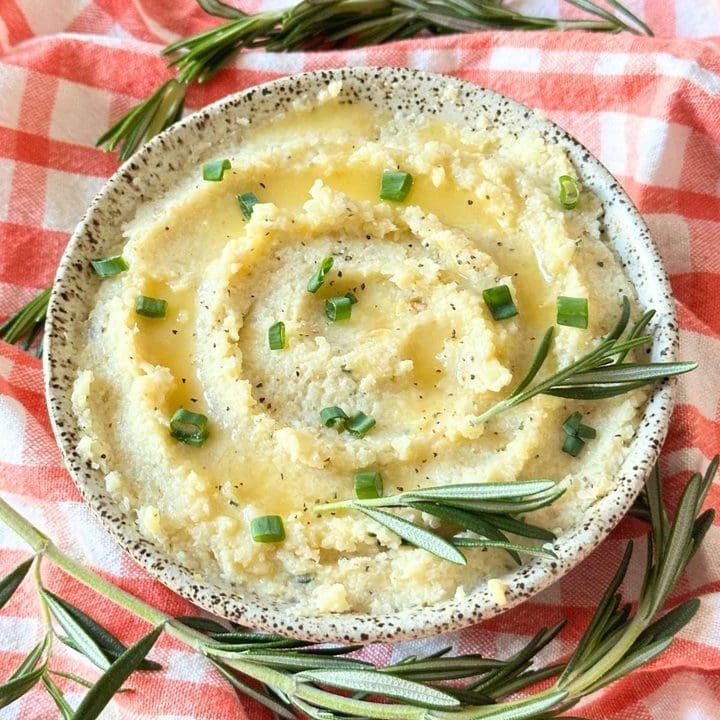Small bowl of vegan cauliflower mash topped with melted vegan butter, green onions, and a sprig of rosemary.