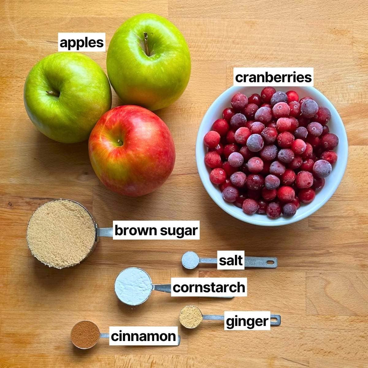 Ingredients to make cranberry apple crisp with corresponding labels: apples (red and green), cranberries, brown sugar, sea salt, cornstarch, ginger, and cinnamon.
