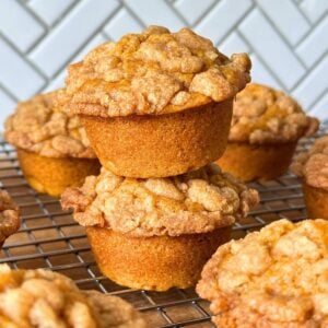 Two vegan pumpkin muffins with a brown sugar streusel stacked on top of one another on a wire rack with more fluffy pumpkin muffins.