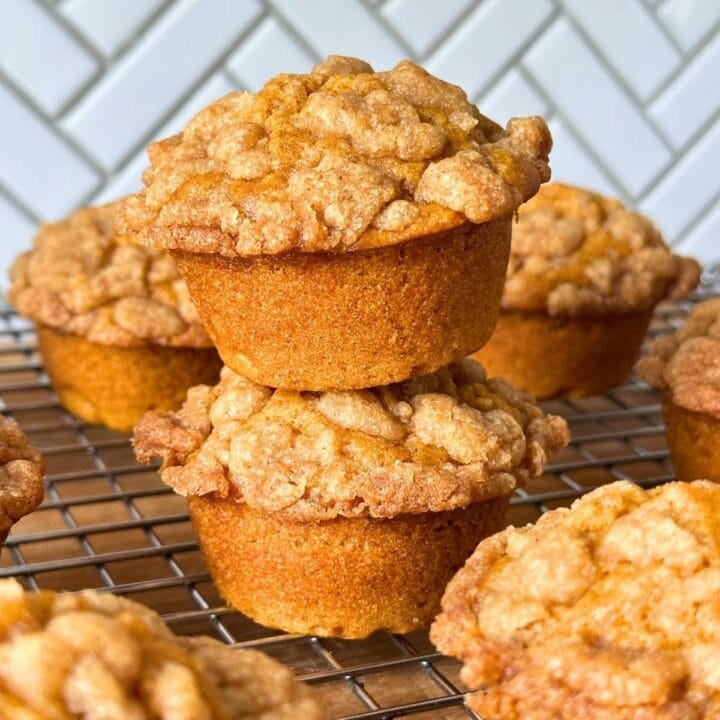 Two vegan pumpkin muffins with a brown sugar streusel stacked on top of one another on a wire rack with more fluffy pumpkin muffins.