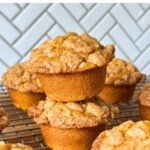 Two vegan pumpkin muffins with streusel stacked on top of one another on a wire rack with more muffins.
