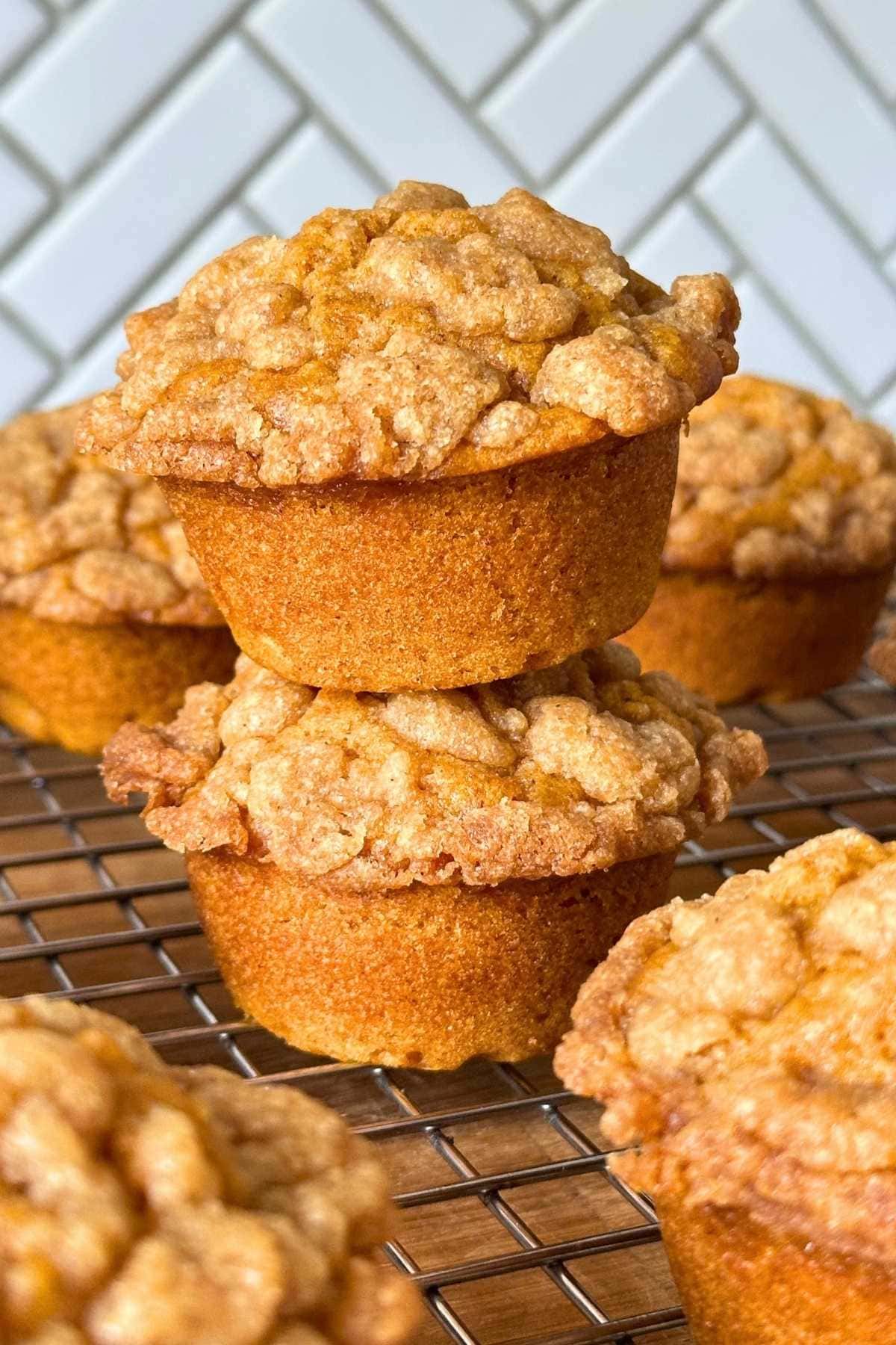 Two vegan pumpkin muffins with a brown sugar streusel stacked on top of one another on a wire rack with more fluffy pumpkin muffins.